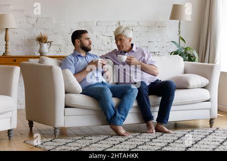 Older man grownup son talking seated on sofa with teacups Stock Photo