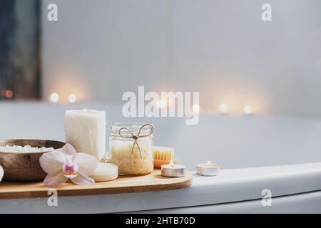 Preparation for hotel spa treatment or home bath procedure. White washbasin in bathroom with accessories on tray. Burning candles, washcloth, soap, foot brush, bottle with sea salt, orchid flower Stock Photo