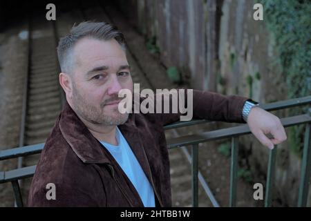Bearded handsome middle aged Caucasian male leaning against the metal fence over the railway tracks Stock Photo