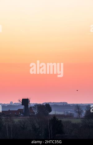 A clear dawn orange sky over a landscape of hedgerows and trees with a English windmill without sails, near the horizon. Stock Photo