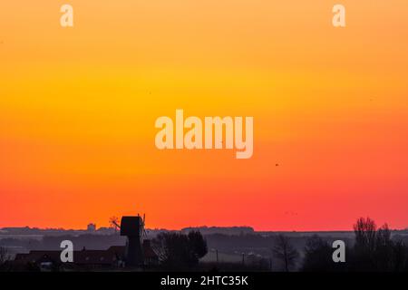A clear dawn orange sky over a landscape of hedgerows and trees with a English windmill without sails, near the horizon. Stock Photo