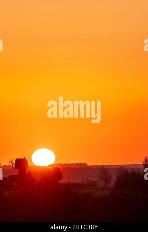 Sunrise in to a clear orange sky over a landscape of hedgerows and trees with a English windmill without sails, near the horizon. Stock Photo
