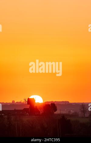 Sunrise in to a clear orange sky over a landscape of hedgerows and trees with a English windmill without sails, near the horizon. Stock Photo