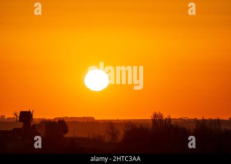Sunrise in to a clear orange sky over a landscape of hedgerows and trees with a English windmill without sails, near the horizon. Stock Photo