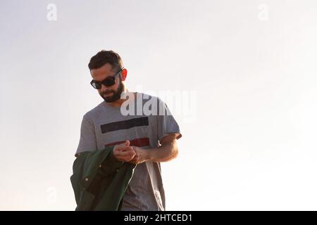 handsome man in sunglasses taking off his jacket with sky in background, concept of leisure and relax, copy space for text Stock Photo