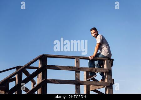 pensive young man sitting on a wooden railing with blue sky in the background, concept of leisure and relax, copy space for text Stock Photo