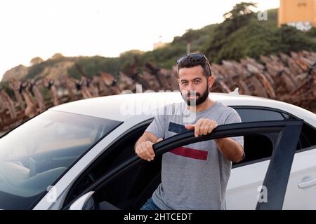 handsome bearded young man looking at camera leaning on the door of his car, road trip and driving concept, copy space for text Stock Photo