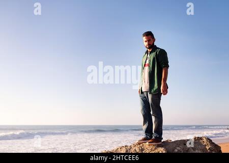 handsome bearded man looking serious at camera standing on a beach rock, concept of leisure and relax, copy space for text Stock Photo