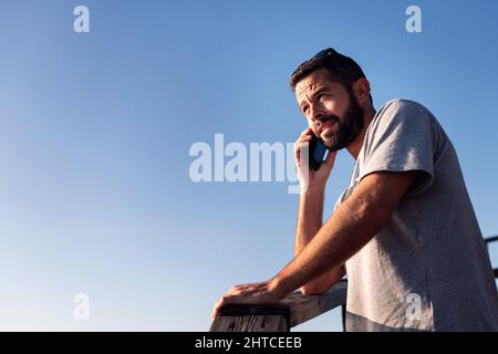 portrait of a handsome young man talking on his smart phone leaning on a wooden railing with blue sky in the background, concept of leisure and relax, Stock Photo