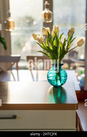 A bouquet of white tulips in a blue green glass vase in bright morning sun. Modern interior design, fresh spring flowers on kitchen counter. Concept o Stock Photo