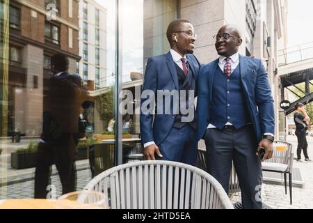 Two dark-skinned African American businessmen friends in suits hug and rejoice at a meeting on the street near a cafe Stock Photo
