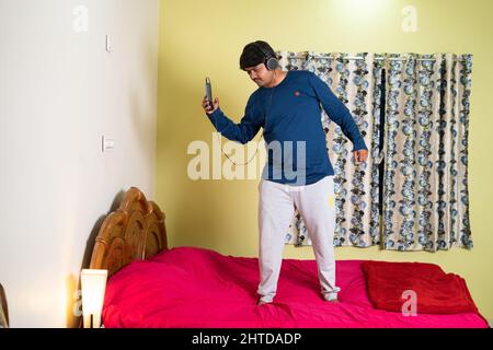 Enjoying young man with headset dancing by singing on bed by listening songs on mobile phone at bedroom - concept of entertainment, leisure activity Stock Photo
