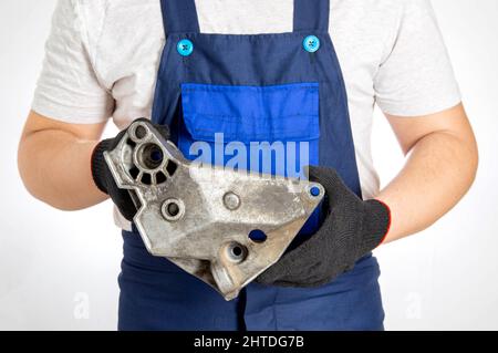 A car mechanic in blue overalls holds a pillow of a car engine in his hands. The concept of replacing an aluminum engine mount in a car, an automotive Stock Photo