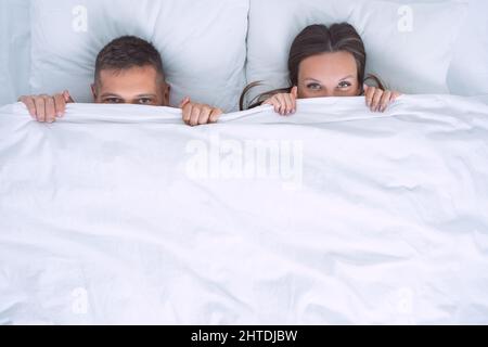 Attractive couple lying in bed and covering face with blanket or hiding under white blanket. Relationship and love concept. Copy space Stock Photo