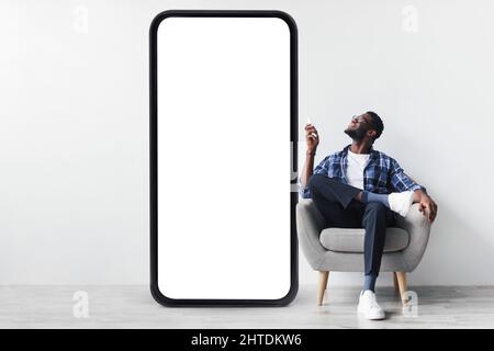 Smart home system. Young black guy with remote control sitting in armchair near huge smartphone, mockup for design Stock Photo