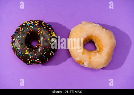 Two Donuts covered with chocolate and sprinkles next to another covered with icing on a purple surface. Stock Photo