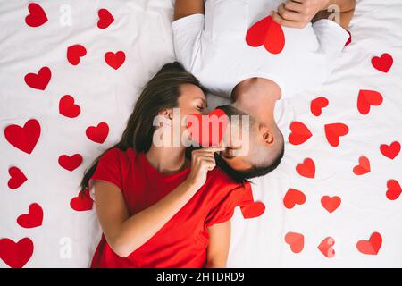 Top view of beautiful couple in love lying on white bed with red hearts and kissing. Love and relationships concept Stock Photo