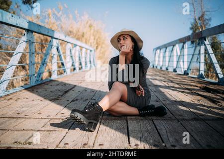 A young Hispanic woman wearing a hat sitting on a dock by the river Stock Photo