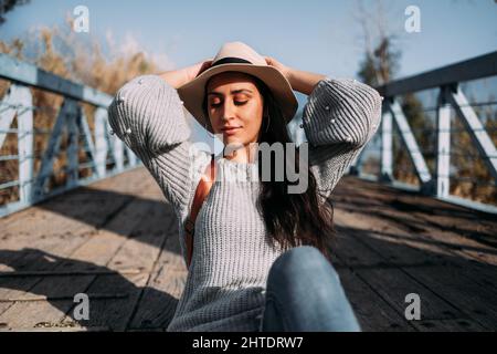 Pretty Hispanic woman wearing a hat relaxing outside in the sun with her arms up Stock Photo