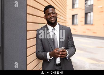 Happy african american entrepreneur in suit using smartphone, standing near office building and smiling at camera Stock Photo