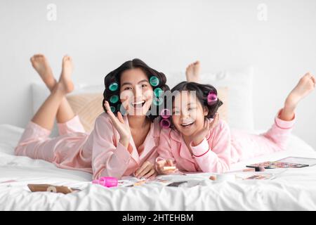 Glad happy millennial japanese lady and teen girl in pajamas and curlers lie on bed with cosmetics, have fun Stock Photo