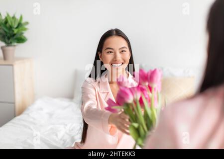 Adolescent girl gives bouquet of tulips, congratulates happy surprised millennial woman in bedroom interior Stock Photo