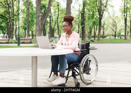 Remote job for people with disability. Young black woman in wheelchair working online, using laptop at outdoor cafe Stock Photo