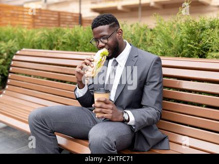 Young african american office worker having lunch outdoors, drinking coffee and eating sandwich, sitting on bench Stock Photo