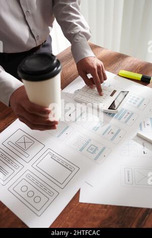 Project manager drinking morning coffee and making calculations after checking mobile app templates Stock Photo