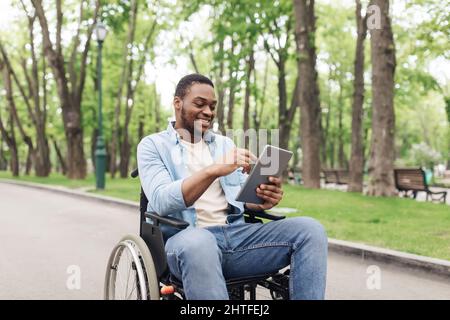 Happy young black man in wheelchair using tablet pc, browsing web or watching movie at spring park Stock Photo