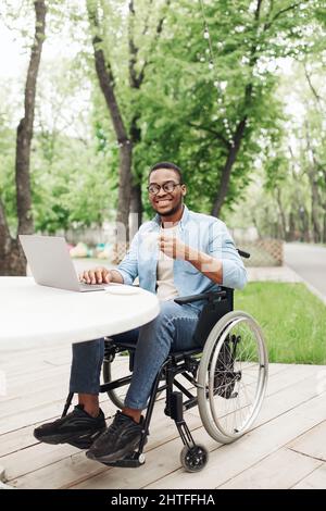 Happy young black man in wheelchair using laptop and drinking coffee at table in outdoor cafe Stock Photo