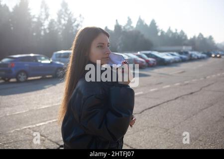 Woman wearing a mask, black jacket and jeans walking in a parking area Stock Photo