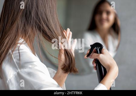 Haircare. Young Female Applying Hair Spray While Standing Near Mirror In Bathroom Stock Photo