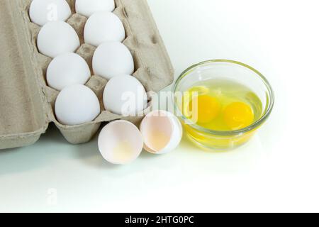 Two broken eggs in a glass bowl with a carton of whole white eggs. Studio shot, white background and copy space. Stock Photo