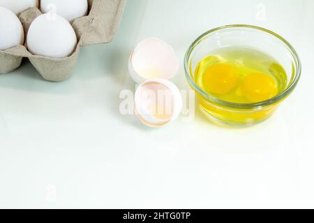 Two broken eggs in a glass bowl with a carton of whole white eggs. White background. Stock Photo