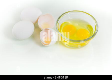 Two fresh whole eggs and two broken eggs in a glass bowl with broken shell near. Studio shot, white background and copy space. Stock Photo