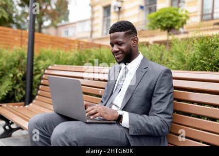 Remote work outdoors. Confident african american businessman or manager in suit using laptop, sitting on bench outside Stock Photo