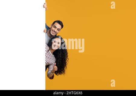 Happy Arabic Man And Woman Peeking Out Behind Big White Advertisement Board Stock Photo