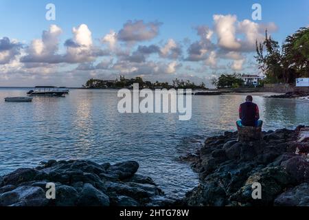 Pereybere beach, Mauritius - Local man man sits on rocks overlooking the beautiful marina in the early morning Stock Photo