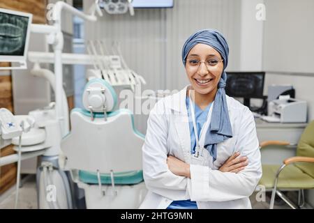 Waist up portrait of young female dentist looking at camera and smiling while wearing headscarf in dental office Stock Photo