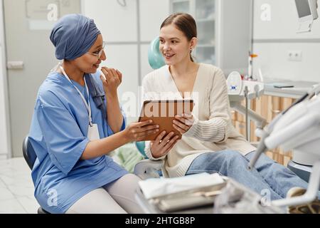 Portrait of smiling young woman in dental chair talking to female doctor and holding digital tablet Stock Photo