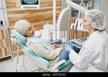 Smiling young woman laying in dental chair and talking to female dentist during consultation in clinic Stock Photo