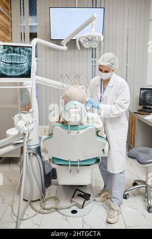 Vertical full length portrait of female dentist wearing full protective uniform examining patient in dental chair at clinic Stock Photo