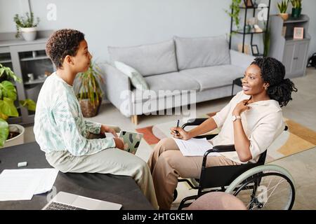 High angle portrait of smiling woman in wheelchair helping daughter doing homework in cozy home interior Stock Photo