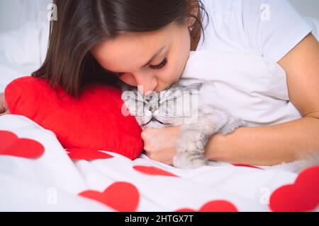 Woman lying on the white bed with red hearts and kissing her cute cat. Cat sleeping with owner. Pets trust and love Stock Photo