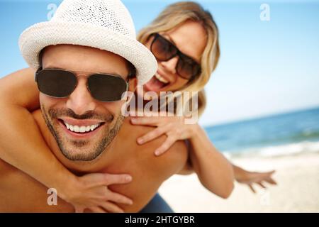 Best vacation EVER. Shot of a smiling young man giving his laughing girlfriend a piggyback on a sunny beach. Stock Photo
