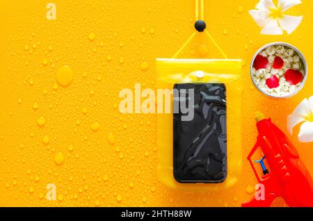 Songkran festival background with waterproof case for smartphone, water gun and flowers in water bowl put on clear mirror that have wet yellow backgro Stock Photo