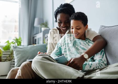 Portrait of happy black mother with daughter doing homework together while sitting on sofa at home Stock Photo