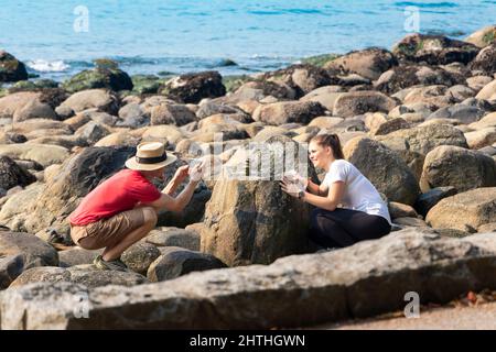 crouching young  man in straw hat takes photograph of female posing on rocky shore in Vancouver canada Stock Photo