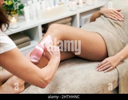 High angle of crop anonymous female master peeling off hair with waxing strip on leg of customer wrapped in towel on couch in professional beauty stud Stock Photo
